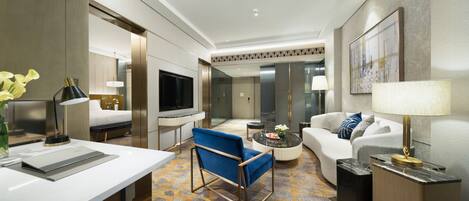 A modern hotel room with a large bed, a blue armchair, a sofa, a TV, and a dining area with a table and chairs.