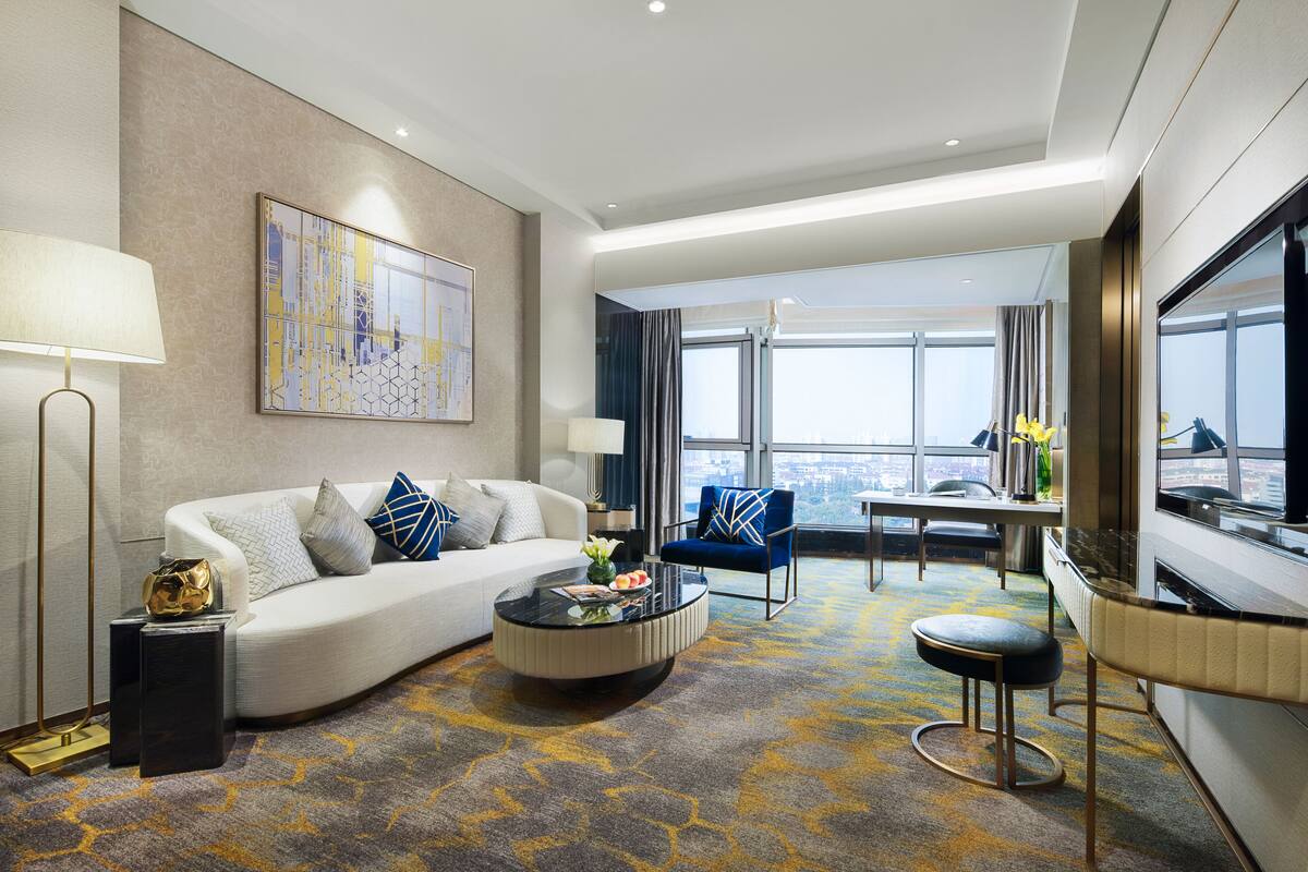 Ein modernes Hotelzimmer mit einer Couch, einem Couchtisch, einem blauen Sessel, einem Essbereich und einem großen Fenster mit Blick auf die Stadt.