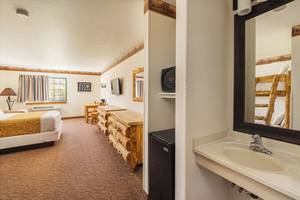 Une chambre d’hôtel comprenant un lit, un banc en bois, un bureau, une télévision et une salle de bain avec un lavabo et un miroir.
