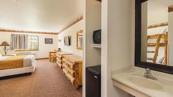 A hotel room with a bed, a wooden bench, a desk, a television, and a bathroom with a sink and mirror.