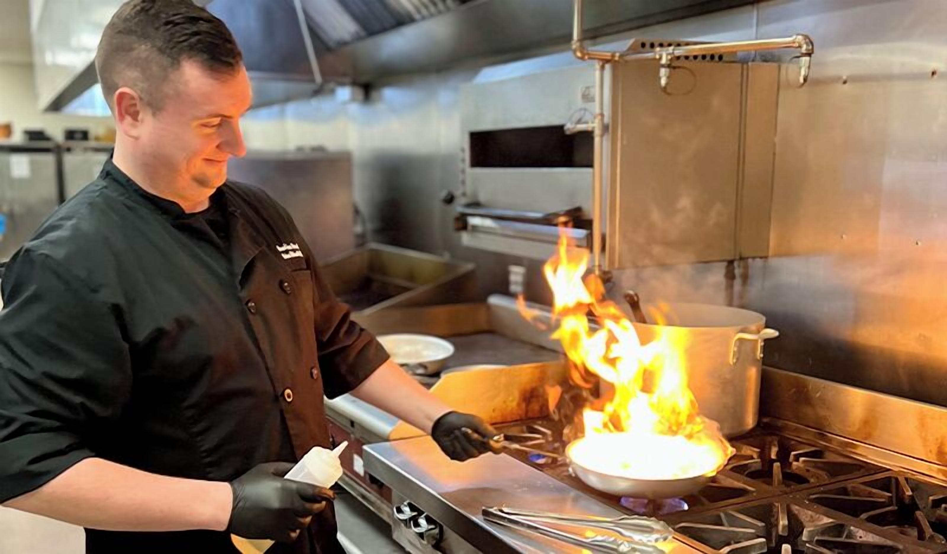 A chef cooking with a pan on a stove with flames.