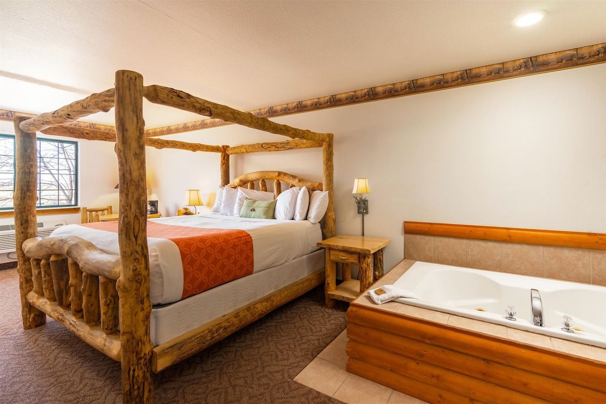 Un lit à baldaquin avec une tête de lit en bois, une table de chevet, une lampe et une grande baignoire.