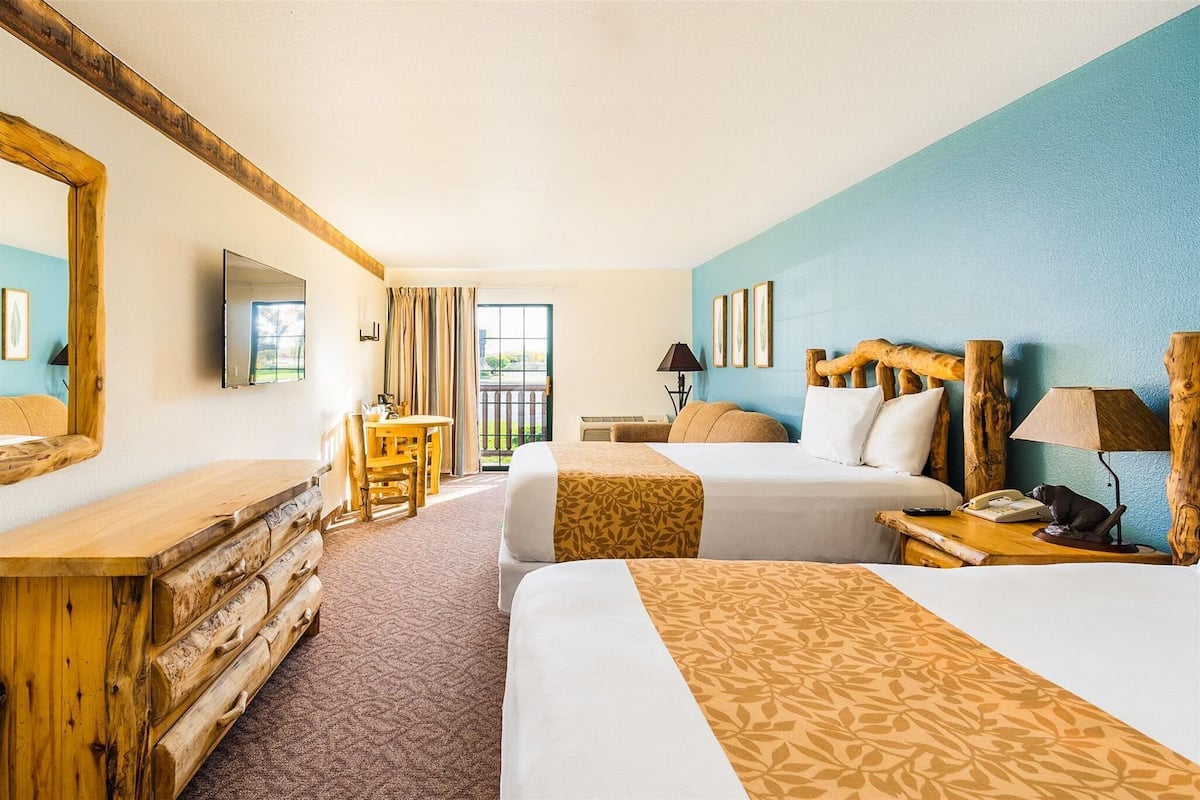 Une chambre d’hôtel avec deux lits, une commode en bois, un bureau avec une chaise, un balcon donnant sur une vue agréable et un grand miroir.