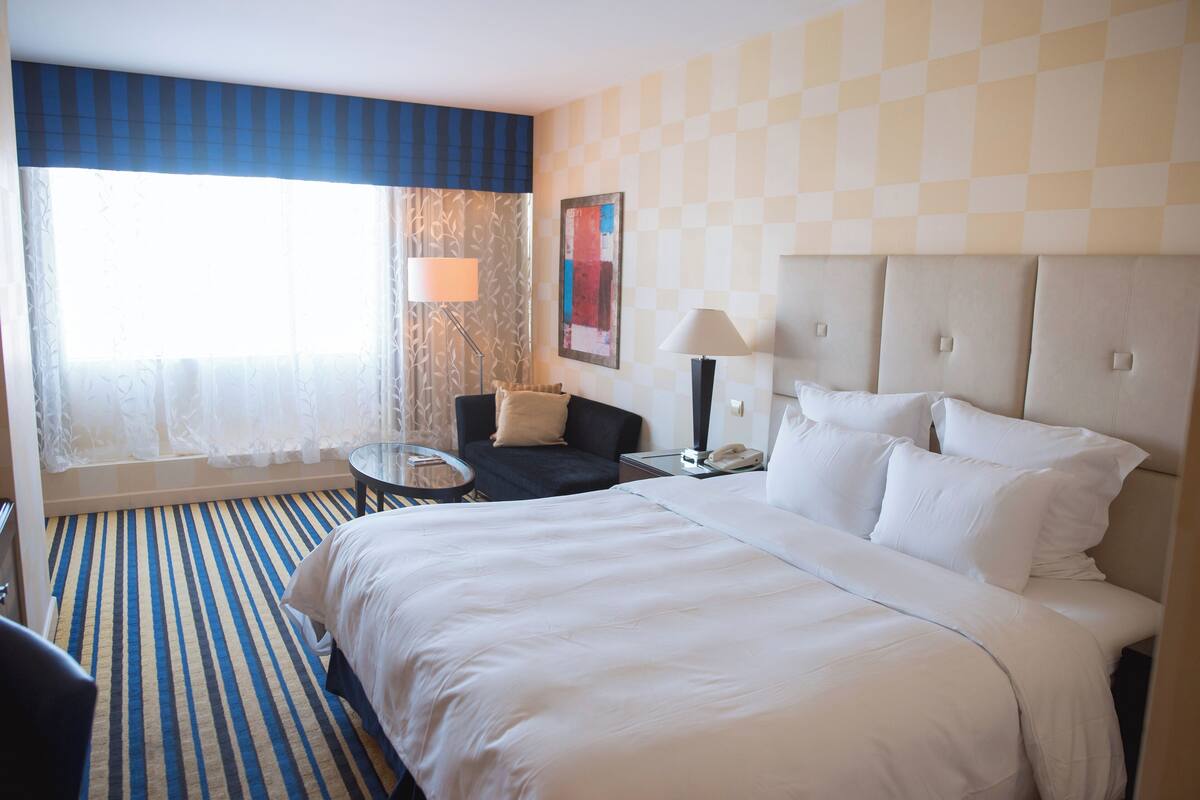 A hotel room with a large bed, a small table, a chair, a lamp, and a window with curtains.