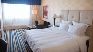 A hotel room with a large bed, a small table, a chair, a lamp, and a window with curtains.