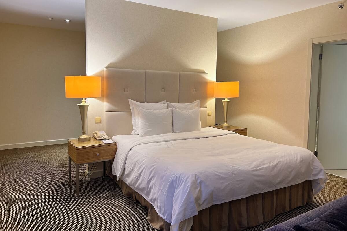 A hotel room with a large bed, two bedside lamps, a nightstand, and a patterned carpet.