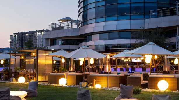 An outdoor seating area with round tables and chairs, umbrellas, and a building with a curved facade in the background.