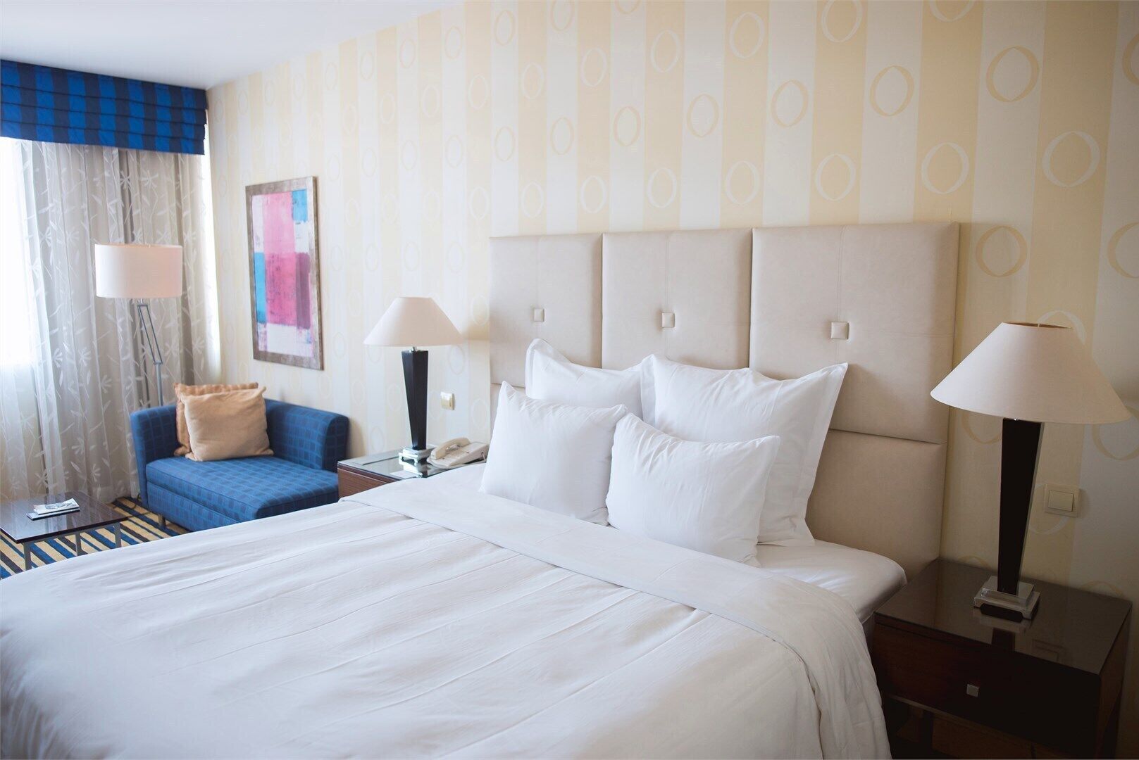 Une chambre d’hôtel avec un grand lit, un fauteuil bleu, deux tables de chevet avec des lampes et un tableau au mur.