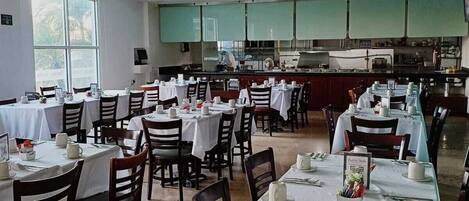 A restaurant with multiple tables set for dining, a kitchen area in the background, and large windows allowing natural light.