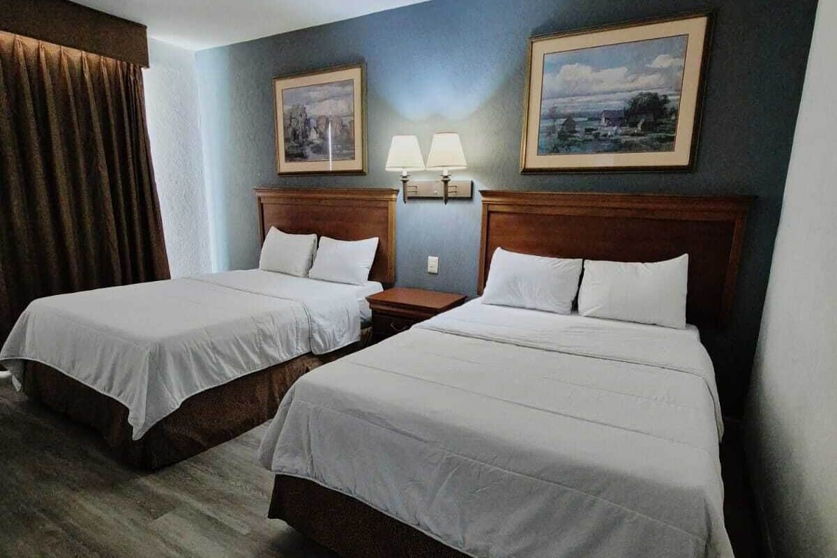 Habitación de hotel con dos camas, cabeceros de madera, una mesita de noche con lámparas y dos cuadros en la pared.