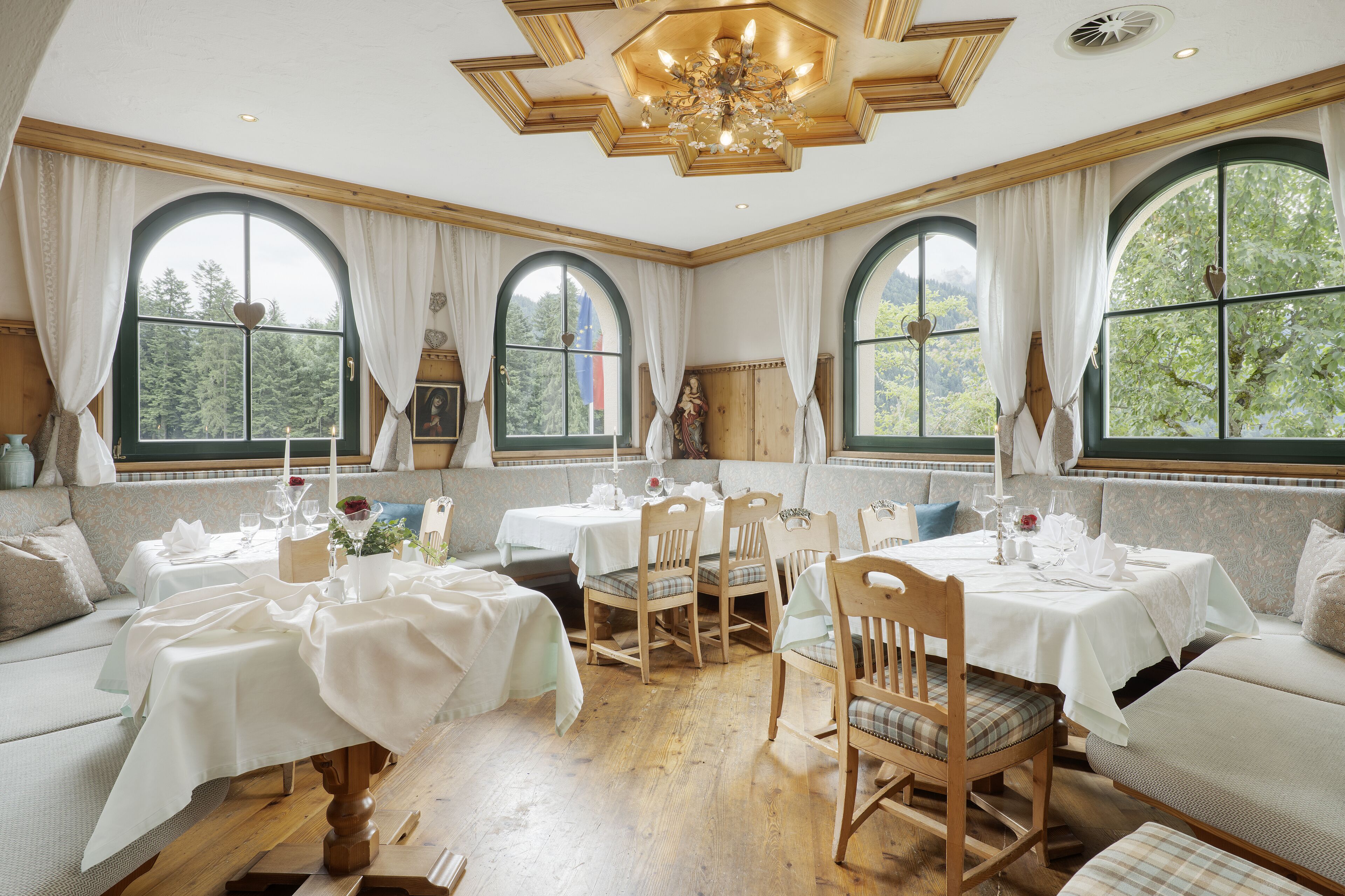 Un espace repas comprenant plusieurs tables et chaises, de grandes fenêtres avec des rideaux et un lustre.