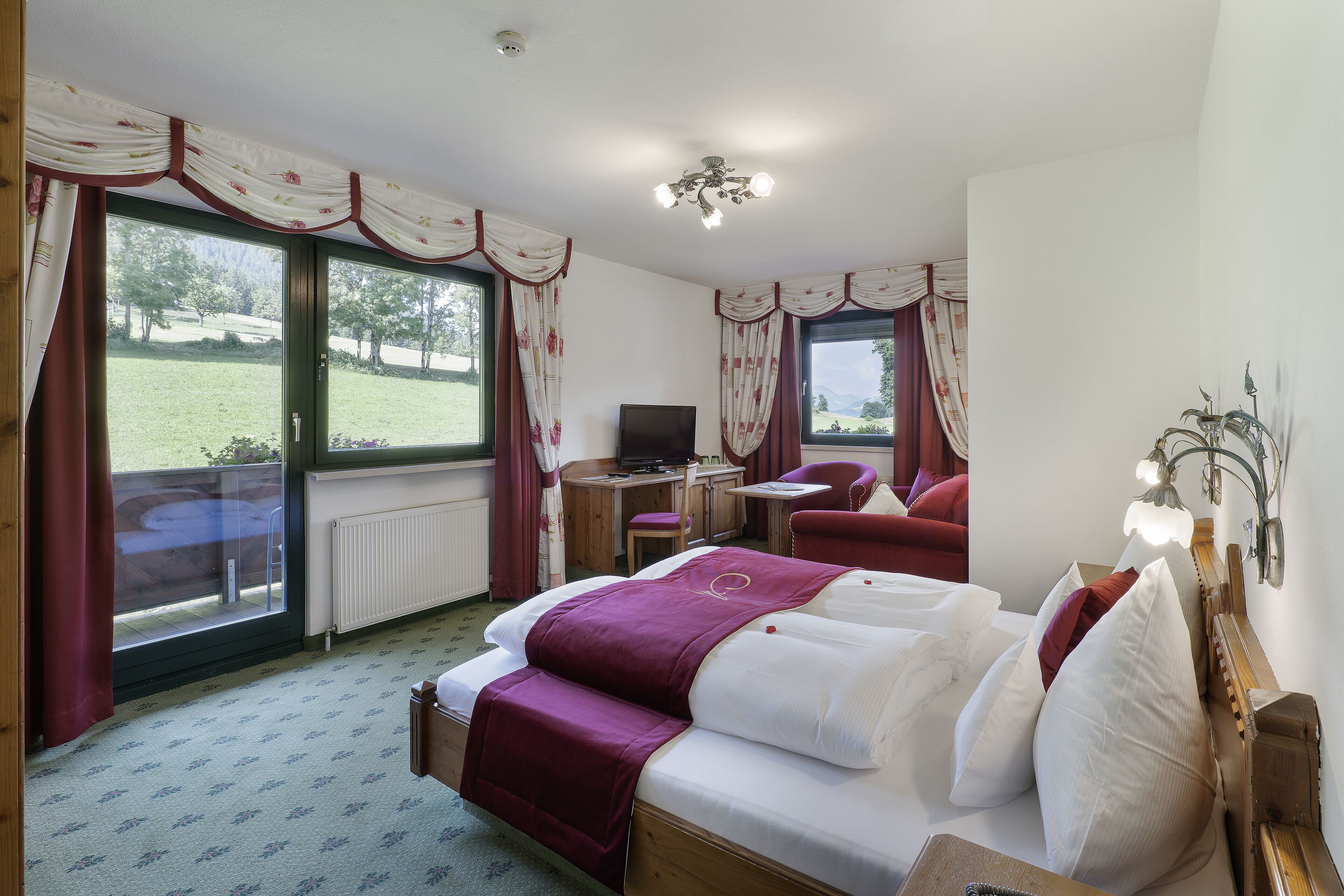 A hotel room with a bed, a desk, a chair, and a view of a grassy area through large windows.