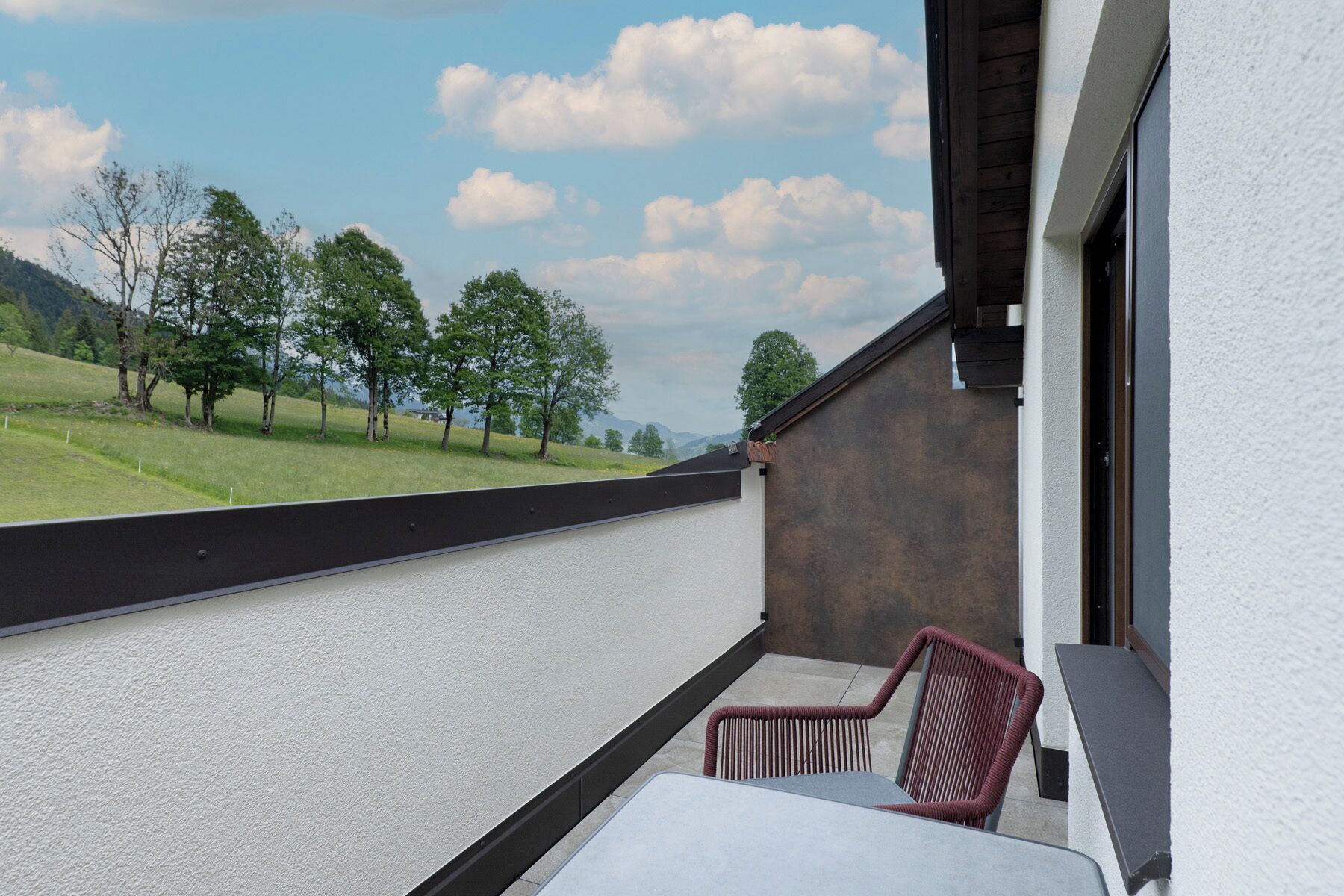 A balcony with a view of a grassy landscape and trees, a chair, and a table.