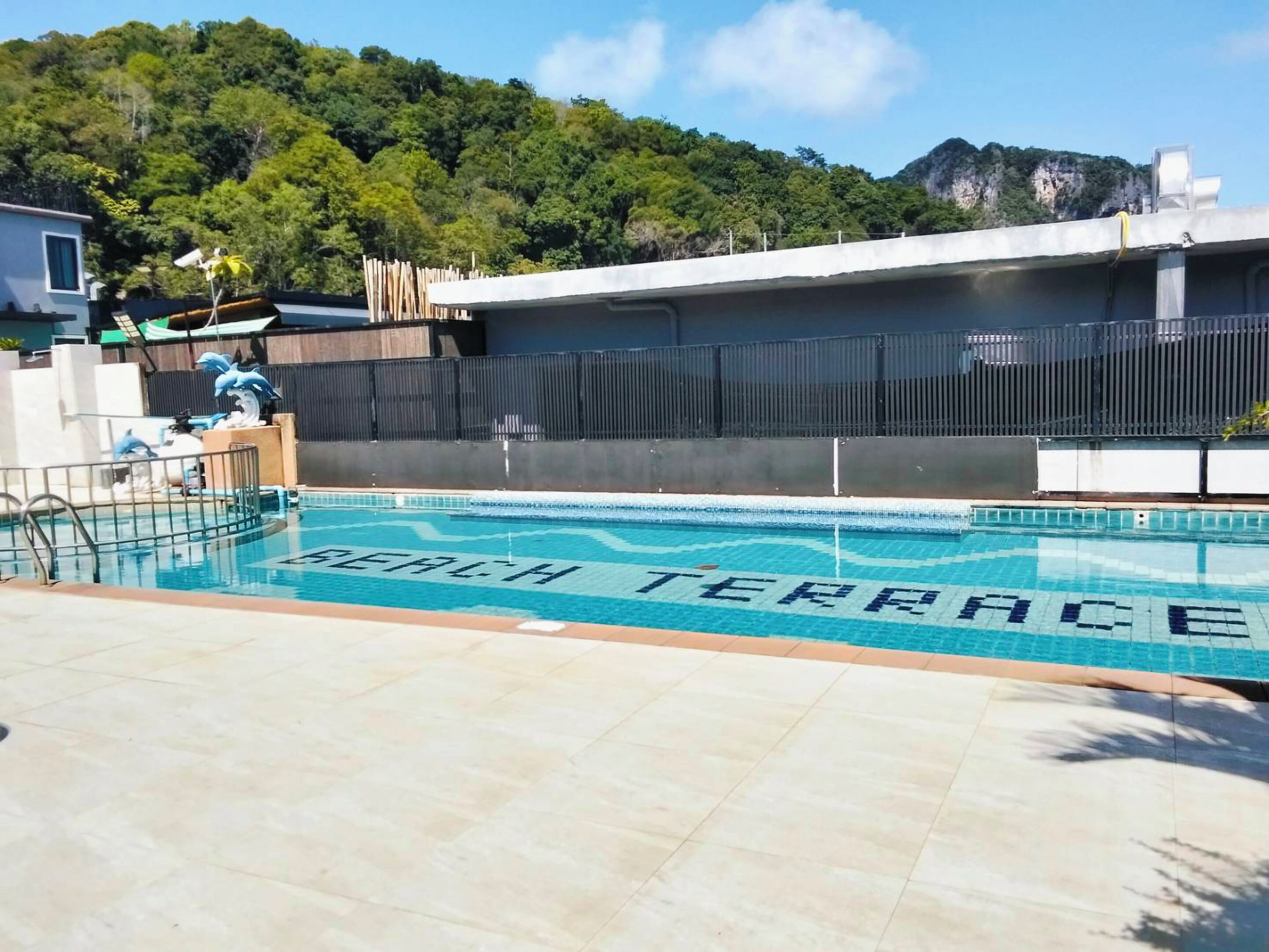 Una piscina con un cartel que dice 'Beach Terrace', rodeada por una valla y con vistas a una colina boscosa.