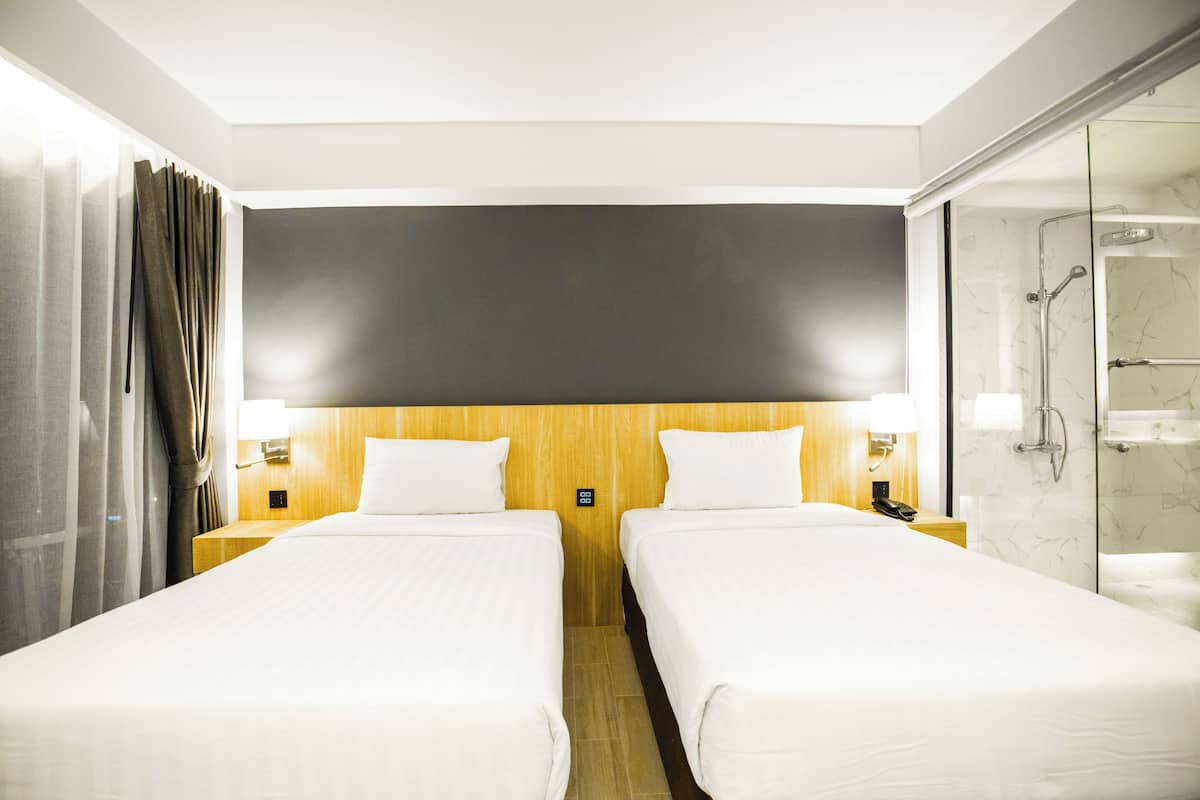 Een hotelkamer met twee enkele bedden, een houten hoofdbord, een douchecabine met glazen wanden en een gordijn aan de linkerkant.