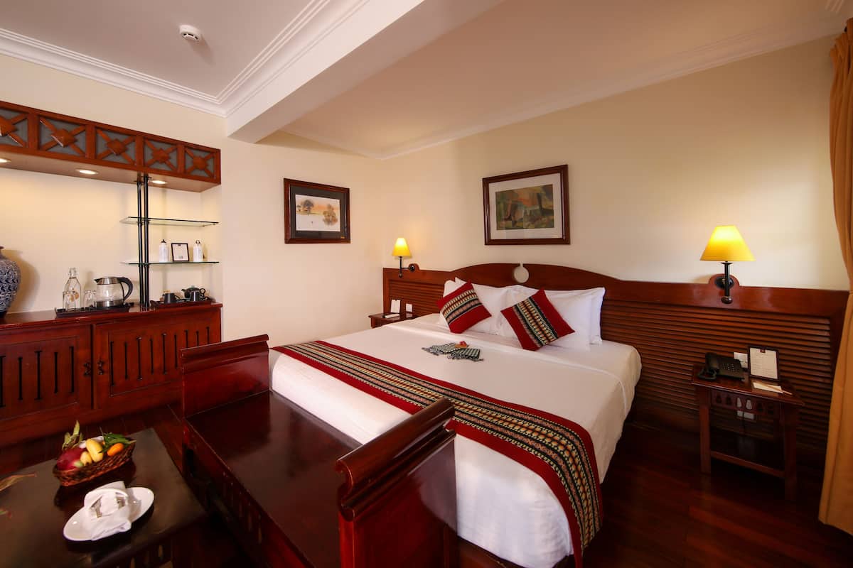 Habitación de hotel con una cama grande, cabecero de madera, dos mesitas de noche con lámparas, cómoda de madera, un espejo y una estantería en la pared con objetos decorativos.