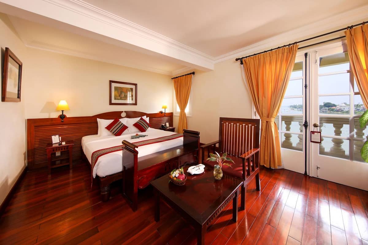 Habitación de hotel con cama de madera, escritorio, silla, mesita de noche, lámpara, cuadro enmarcado y vista a un cuerpo de agua a través de una puerta de cristal.