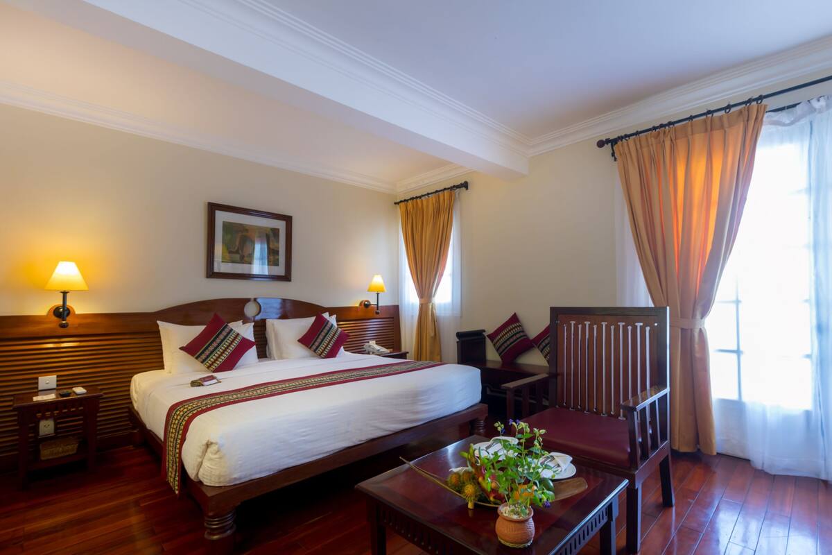 Una habitación de hotel con una cama grande, mesitas de noche, una silla y una pequeña mesa con una planta.