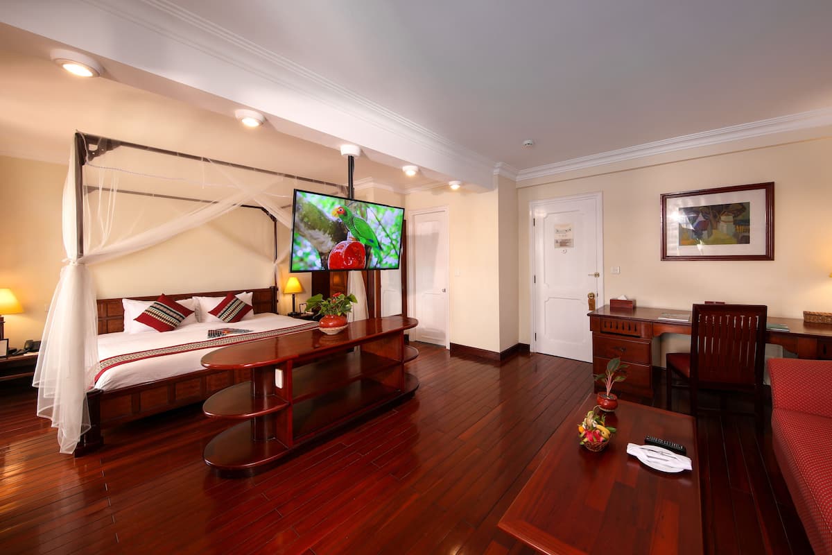 Habitación de hotel con cama con dosel, un escritorio con lámpara, un televisor y una puerta.