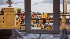 Comedor con vista a un cuerpo de agua, con una mesa servida, platos, vasos y una planta en maceta.