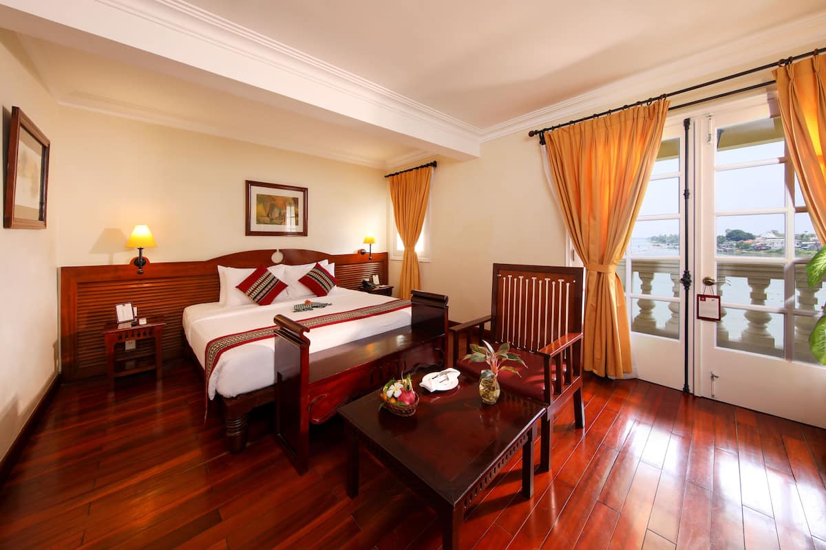 Habitación de hotel con una cama de madera, una mesita de noche con lámpara, una silla, una pequeña mesa con un jarrón y vistas al mar a través de un ventanal grande.
