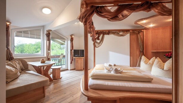 Un dormitorio con cama con dosel, un banco de madera, una mesita y un televisor en una esquina.