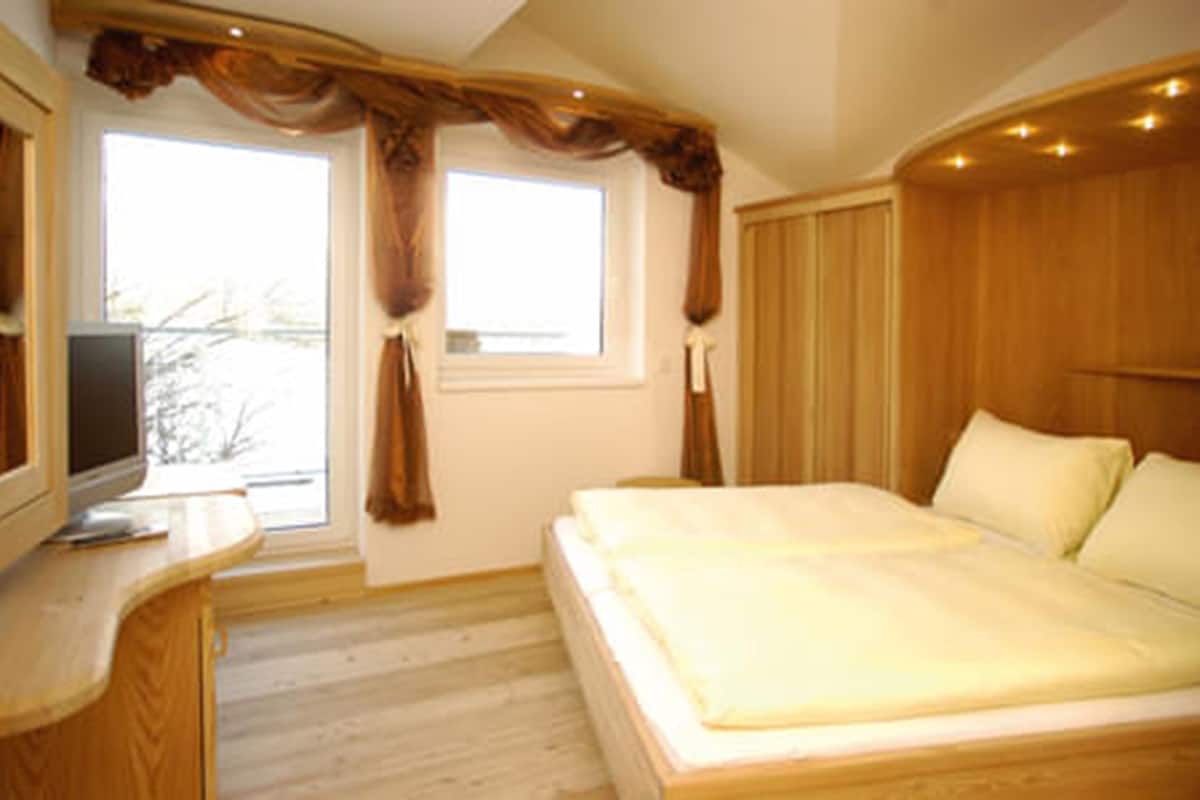 Ein Schlafzimmer mit einem Bett, einem Fernseher auf einer Holzkommode und einem Fenster mit Vorhängen.