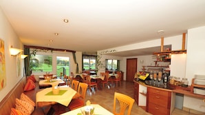 Un comedor con varias mesas y sillas, una zona de cocina con mostrador y estanterías, y ventanas amplias con vistas al exterior.
