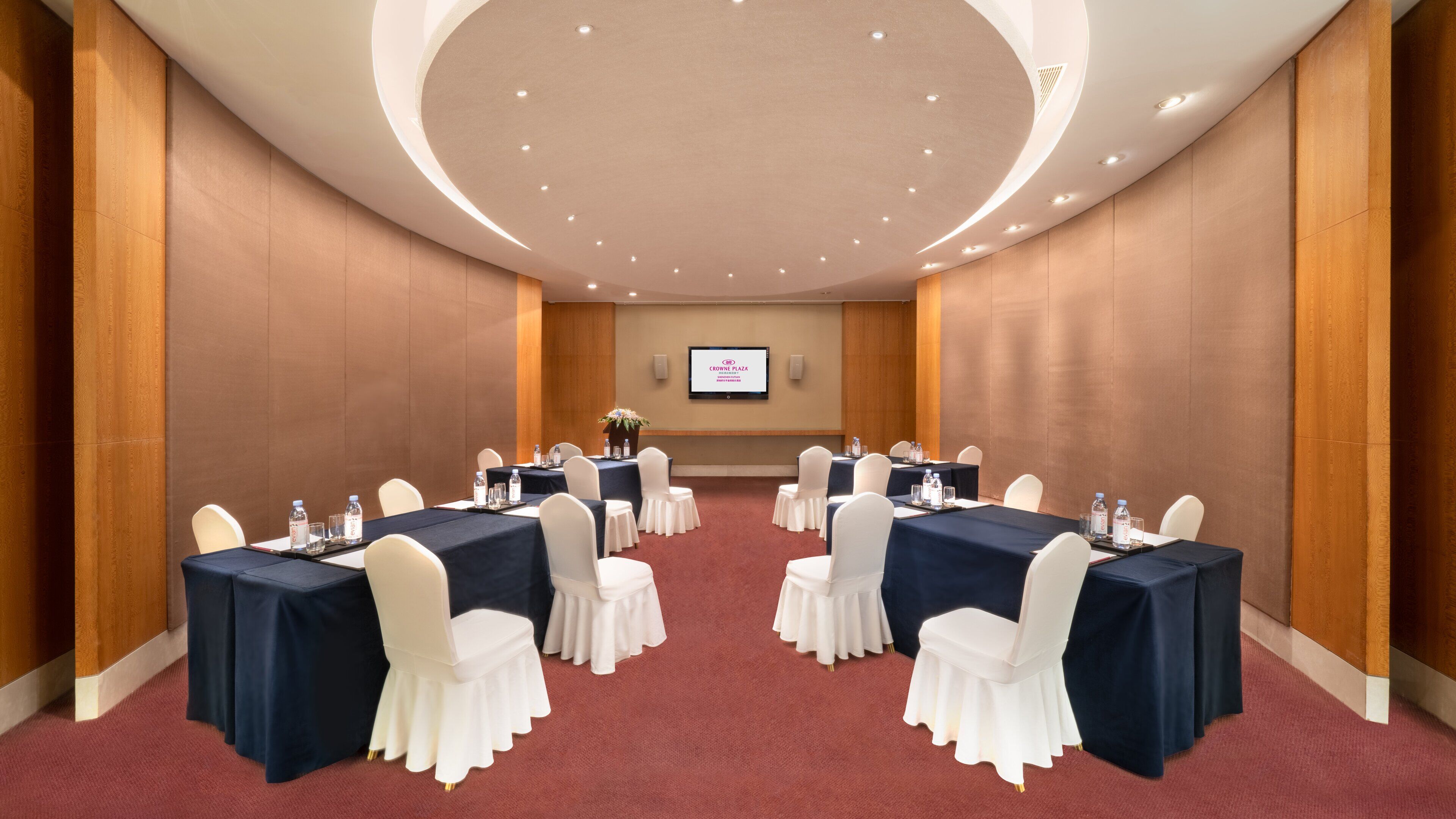 A conference room with tables and chairs, a screen displaying 'Crowne Plaza', and a red carpeted floor.