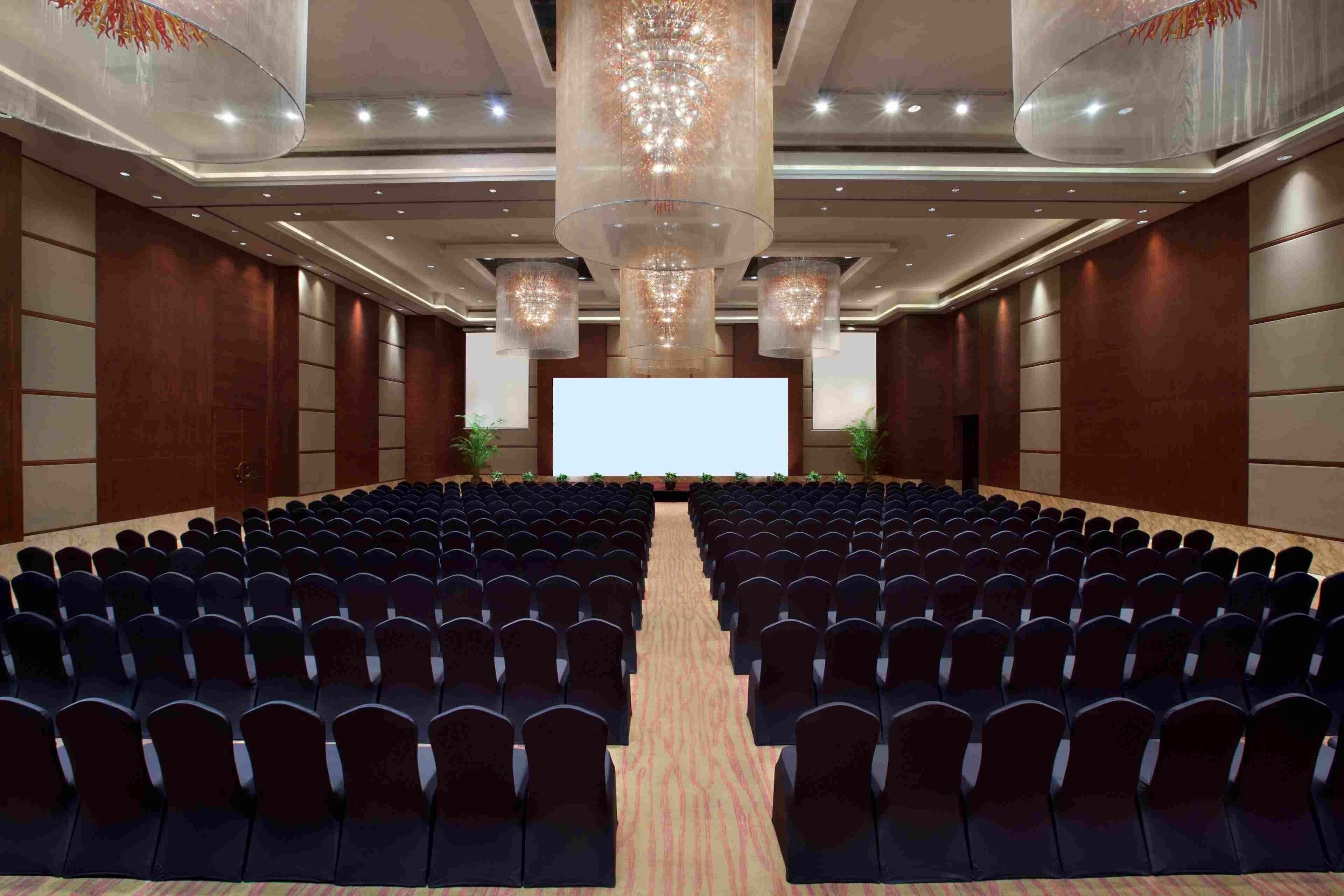 A conference room with rows of chairs facing a large screen, overhead lighting, and a wooden floor.