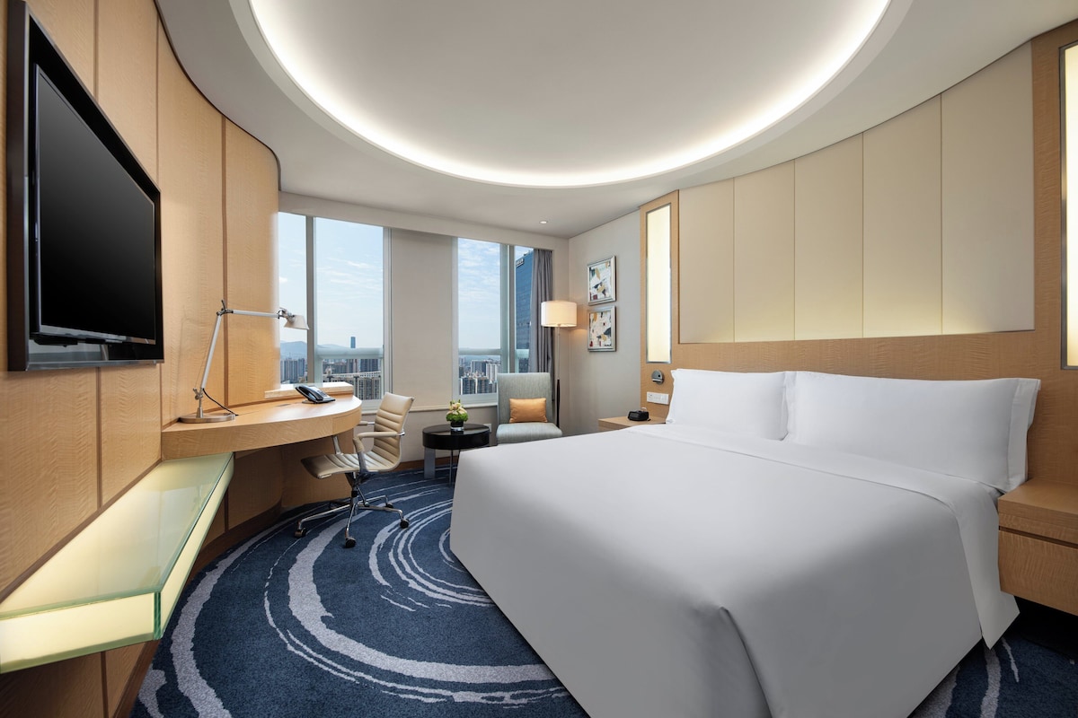 Habitación de hotel moderna con una cama grande, un escritorio con computadora, un televisor de pantalla plana y vistas a la ciudad.