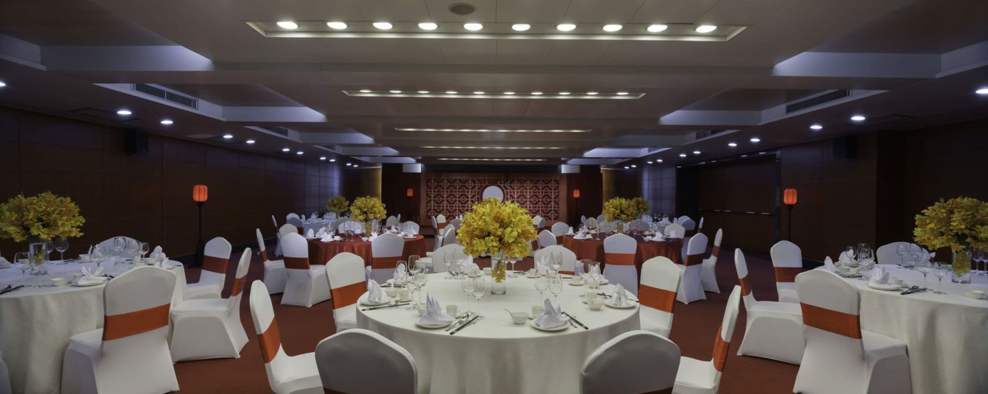 Une salle de banquet avec des tables rondes, des chaises blanches ornées de rubans orange et des centres de table floraux jaunes.