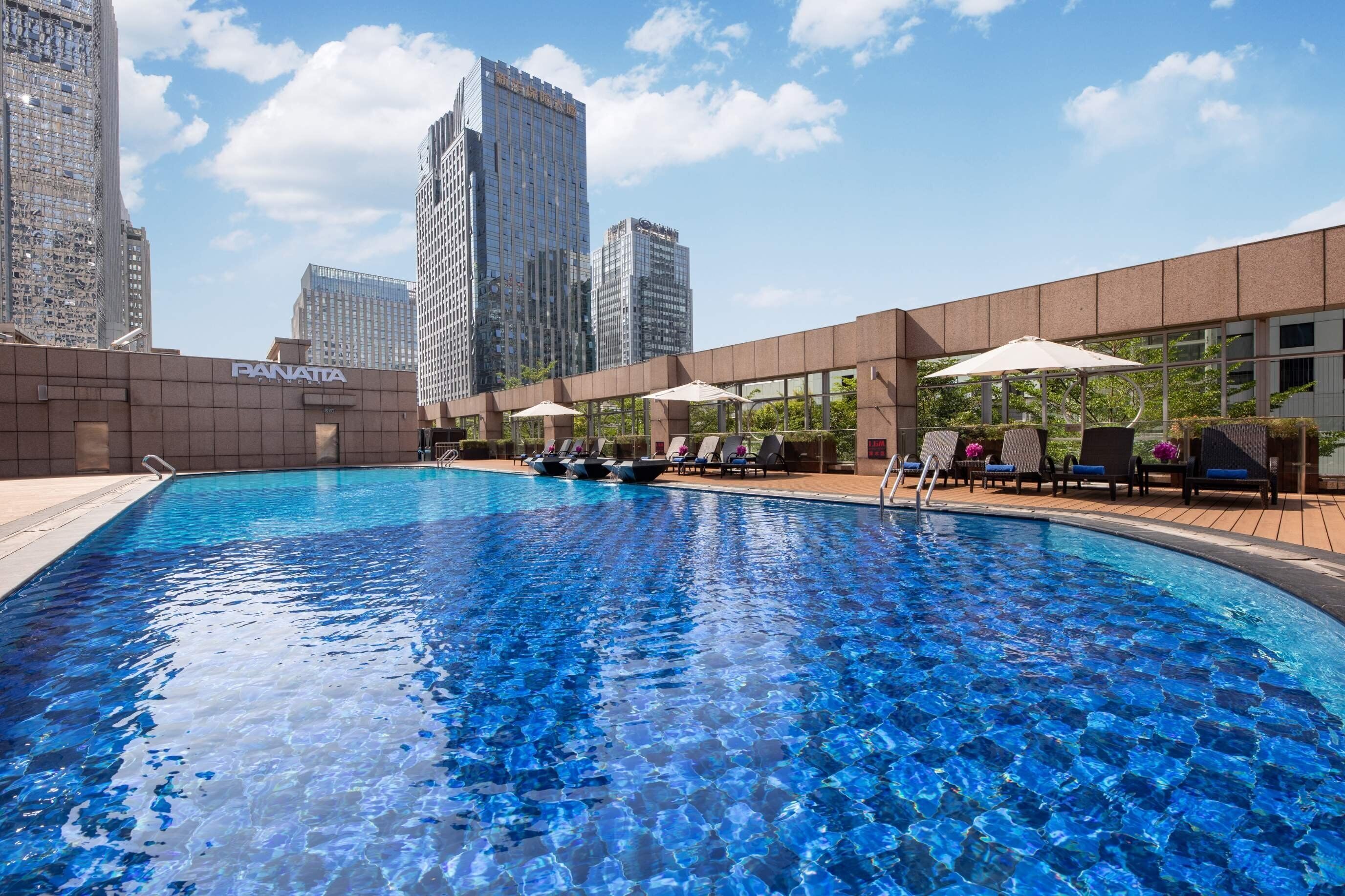 Una piscina con sillas de descanso y sombrillas, rodeada de edificios modernos.