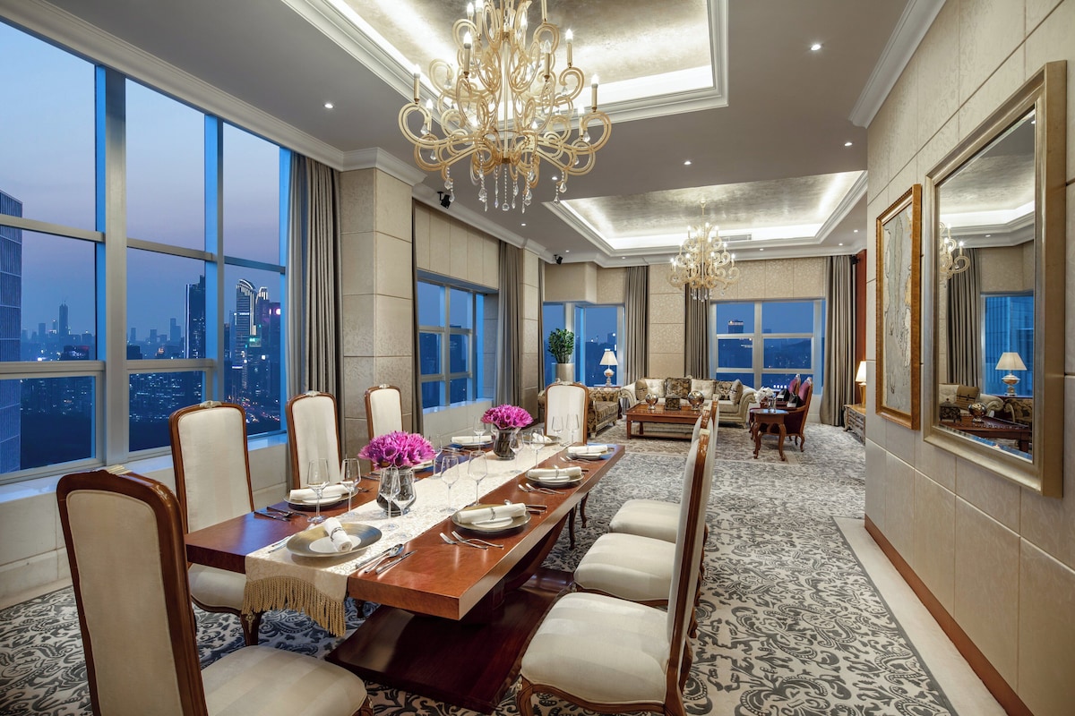 Un comedor amplio con una larga mesa dispuesta para una comida, ventanas grandes que ofrecen vistas de la ciudad y elegantes candelabros.