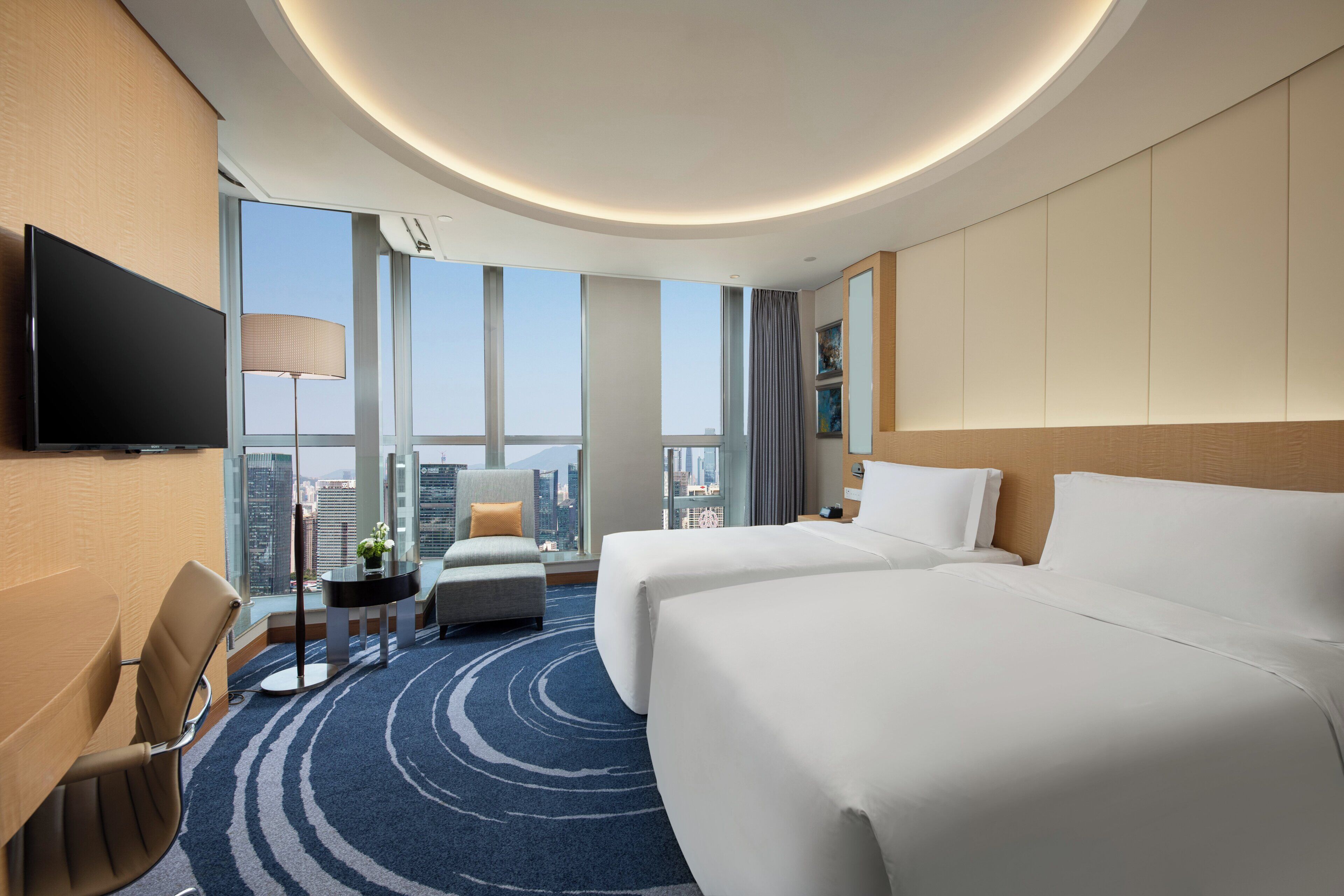 A hotel room with two beds, a TV, a desk, and a view of the city.