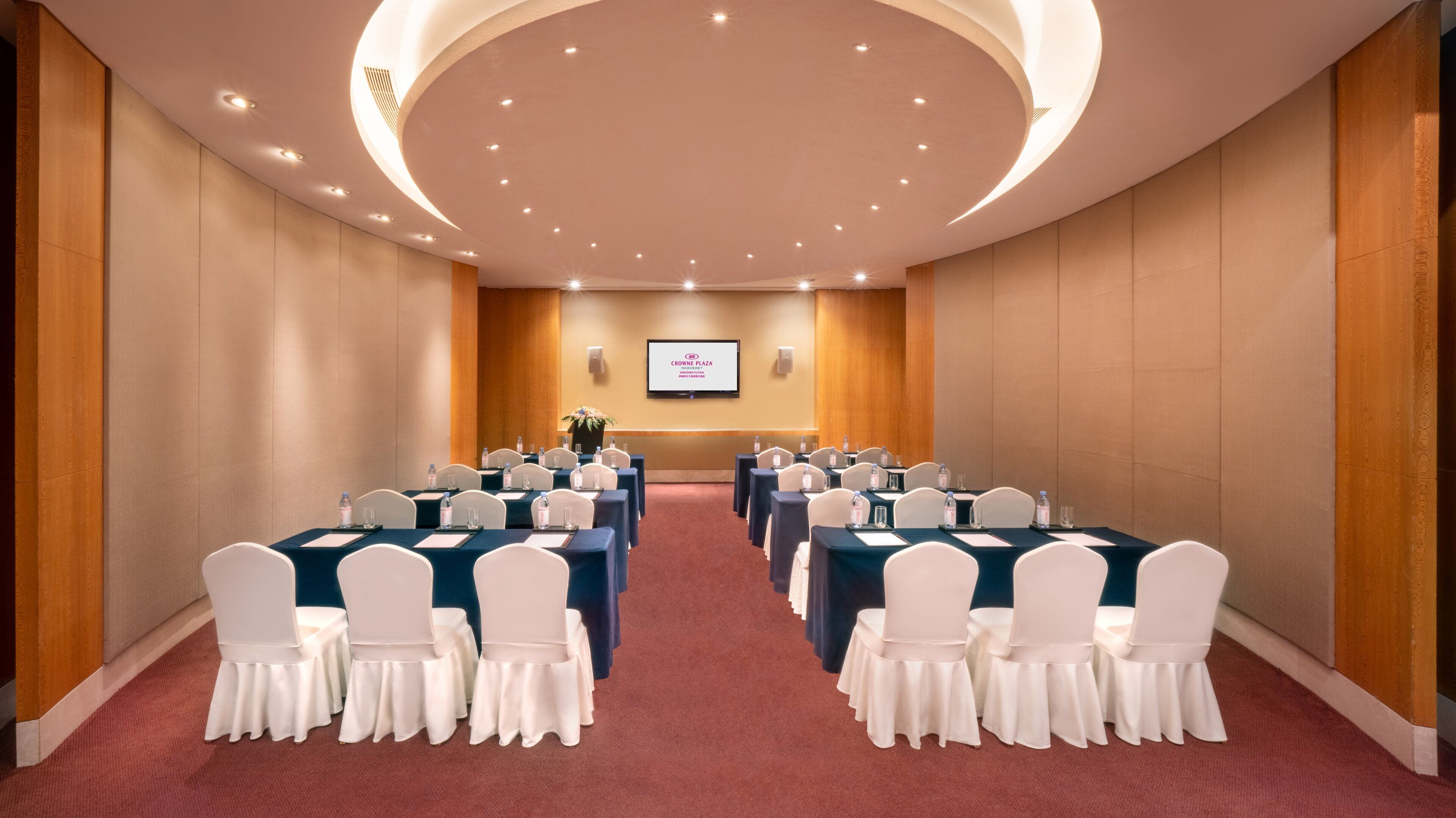 Un salón de conferencias con mesas y sillas, una pantalla mostrando texto, y suelo alfombrado.