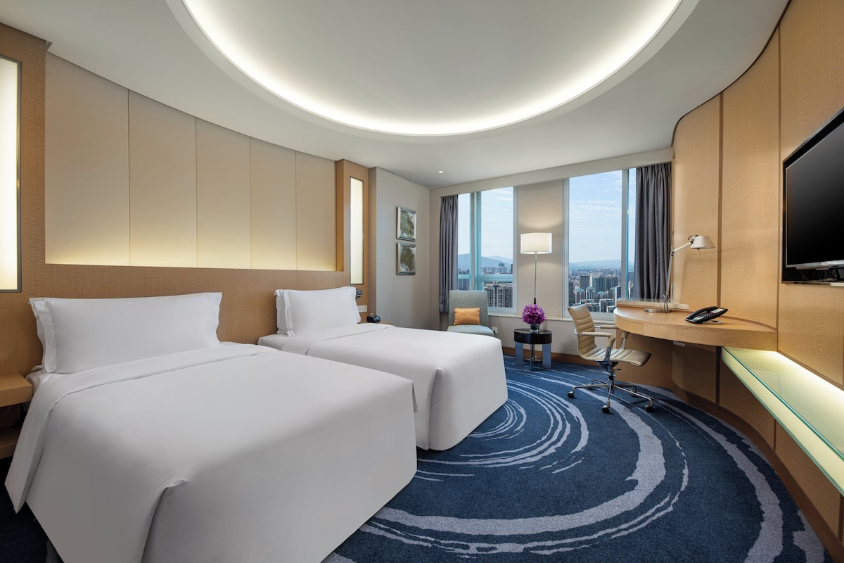 Habitación de hotel con dos camas, un escritorio, una silla, un televisor y vistas a la ciudad.