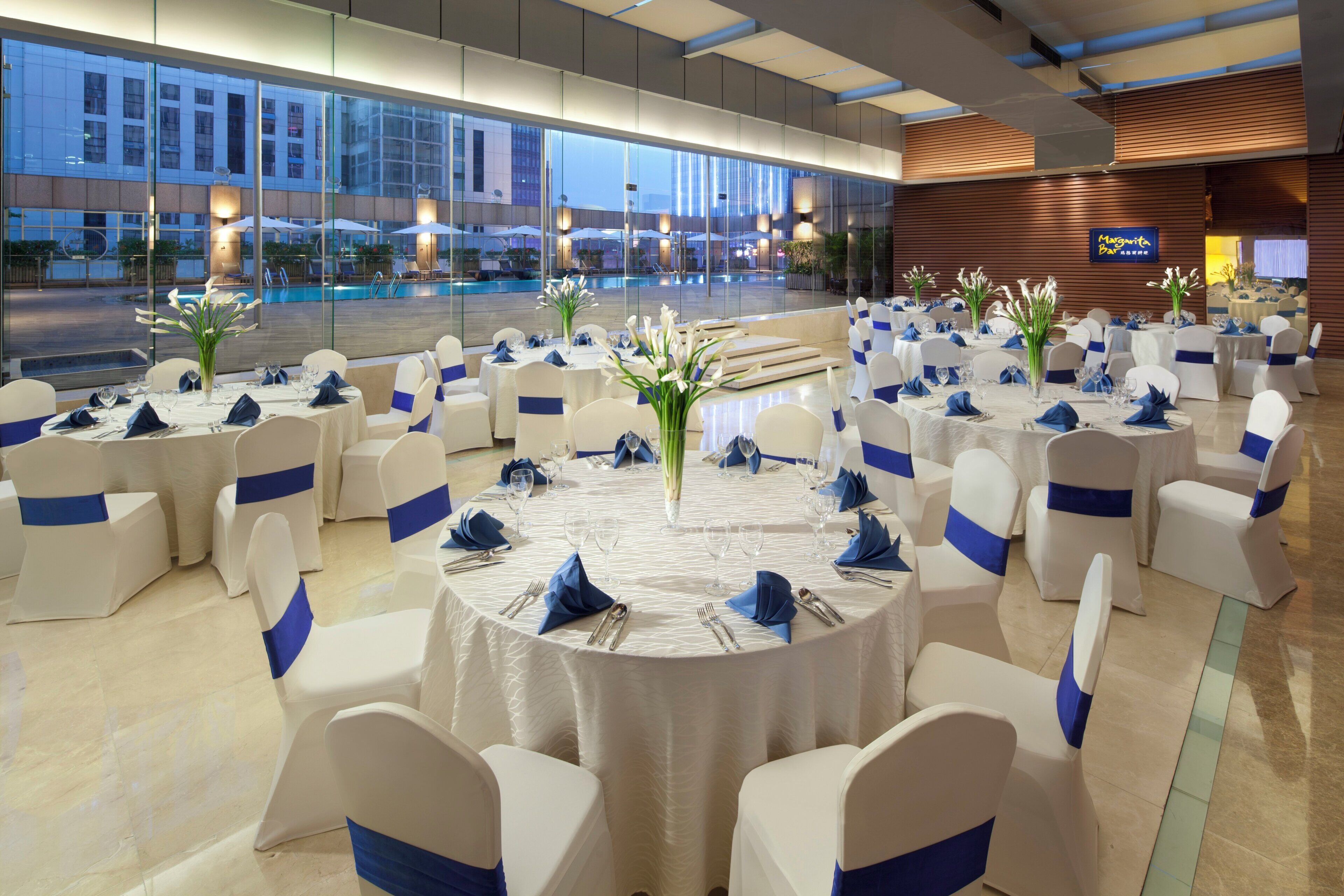Un salón de banquetes con mesas redondas, sillas blancas con cintas azules, y una vista de una piscina y un edificio al fondo.