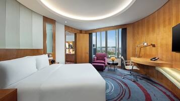 A hotel room with a large bed, a desk with a chair, a small table with a lamp, and a large window offering a city view.