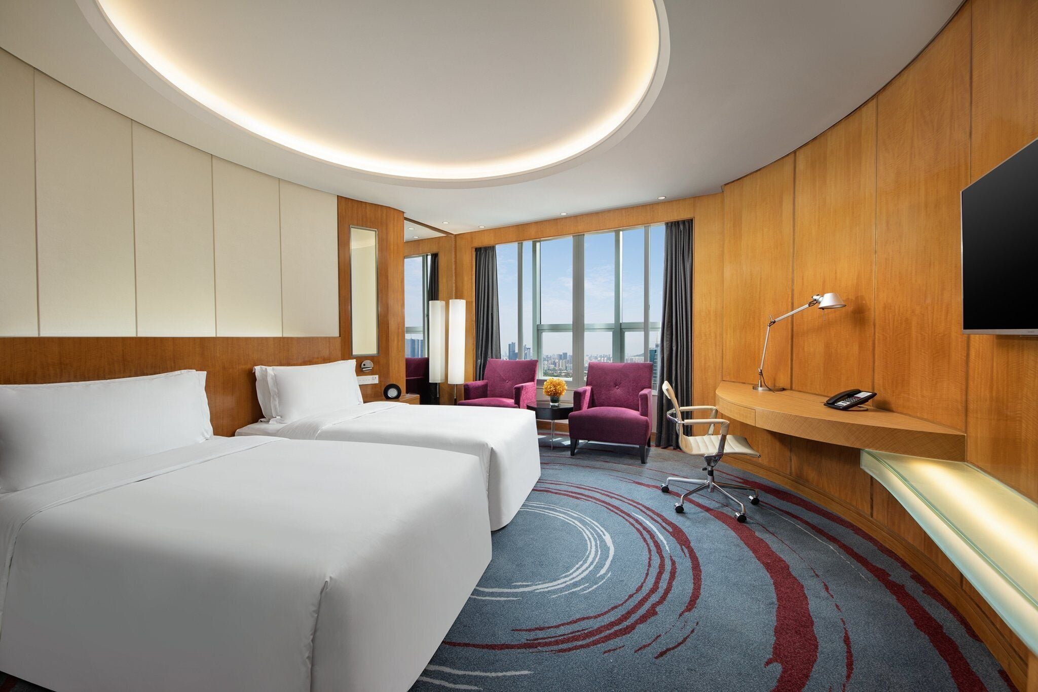 A hotel room with two beds, a desk, a chair, a TV, and a large window with a city view.
