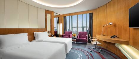 A hotel room with two beds, a desk, a chair, a TV, and a large window with a city view.