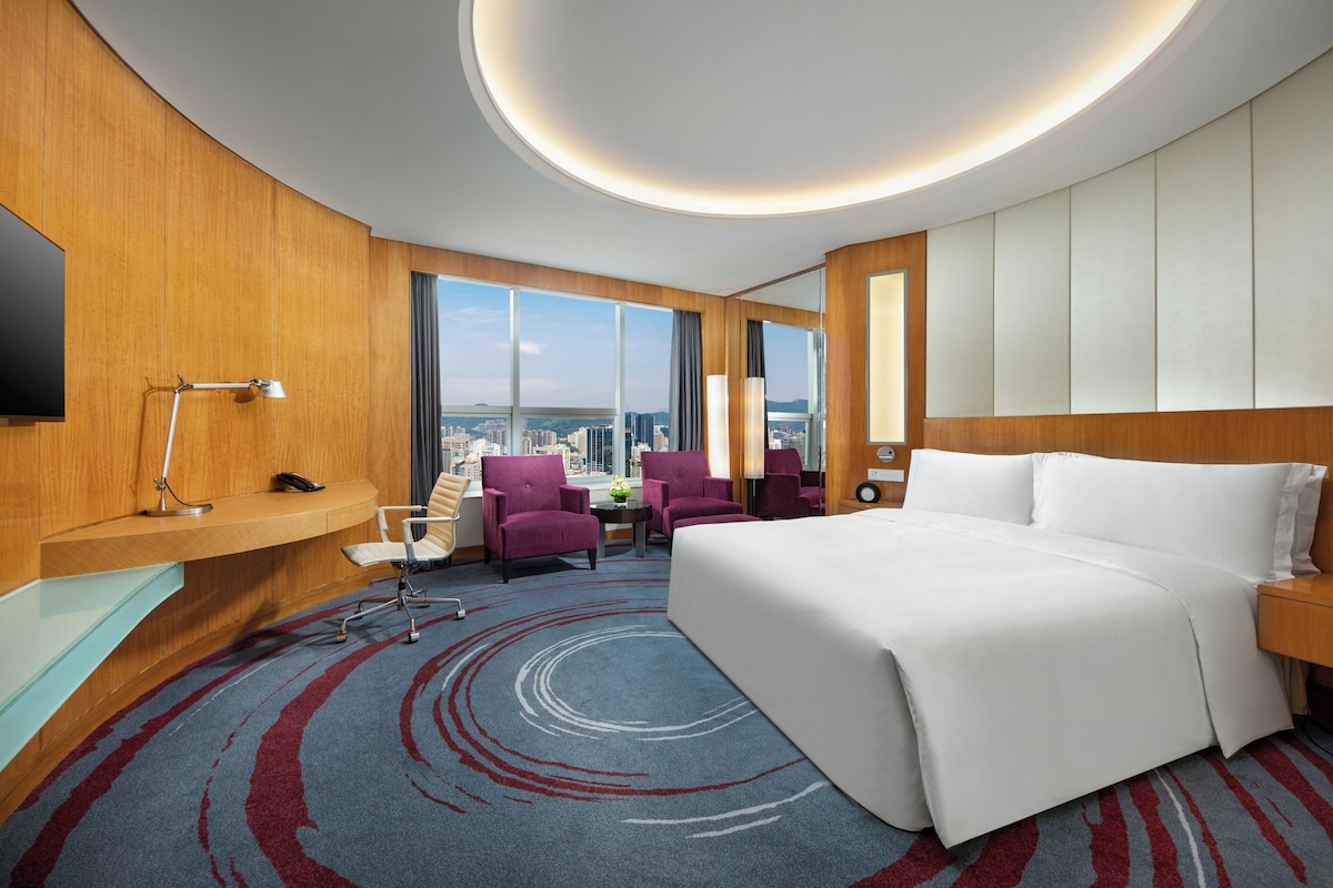 Habitación de hotel con una cama grande, un escritorio con lámpara, una silla, una mesita de noche con un florero y ventanas amplias con vistas a la ciudad.