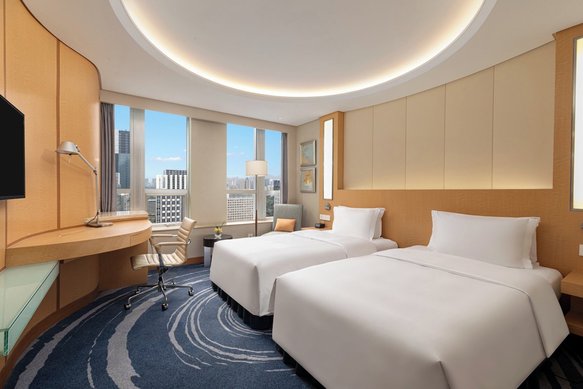 Habitación de hotel con dos camas, un escritorio, una silla, un televisor y vista a la ciudad.