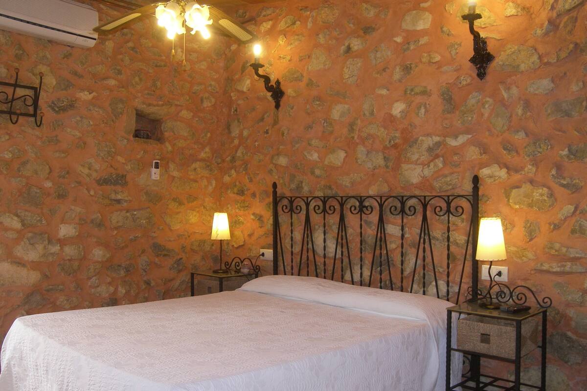 A bedroom with a stone wall, a bed with a white bedspread, two bedside tables with lamps, and a chandelier.