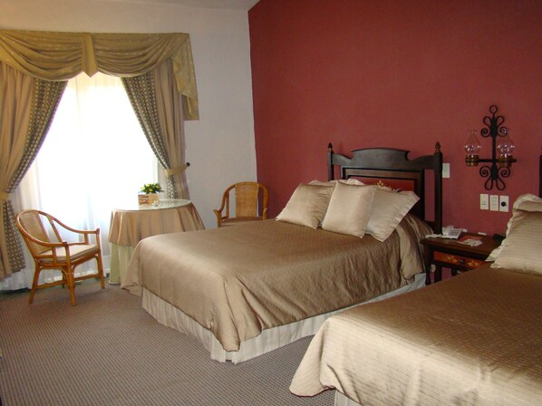 A hotel room with a bed, two chairs, a small table with a lamp, and a wall-mounted light fixture. - Mansión del Cupatitzio (Uruapan)