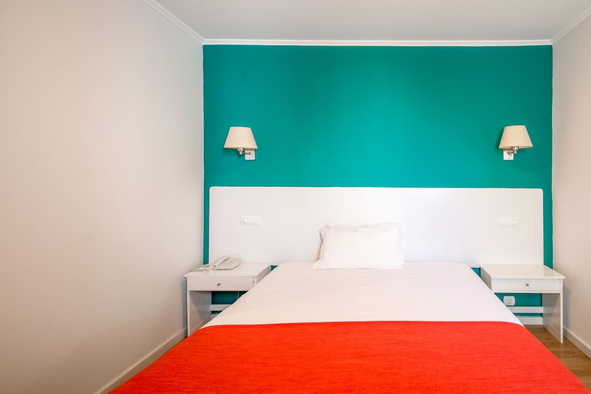 A hotel room with a teal accent wall, white headboard, and a red accent on the bed.