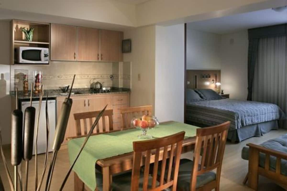 A compact hotel room with a kitchenette, a dining area with a table and chairs, and a bed with a blue blanket.