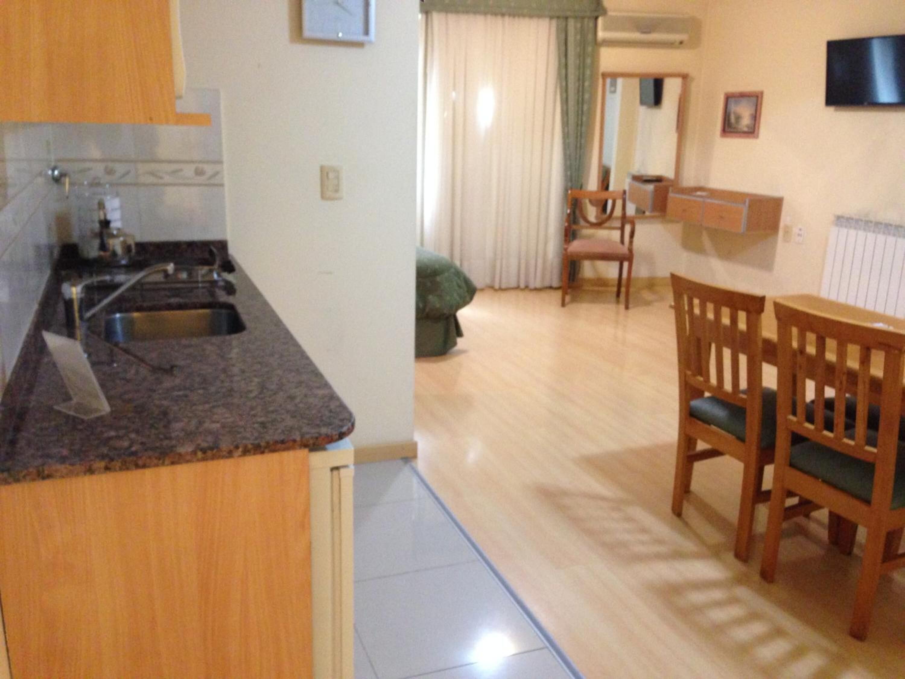 A kitchen with a granite countertop, wooden cabinets, and a sink. There is a dining area with a wooden table and chairs, and a television mounted on the wall.