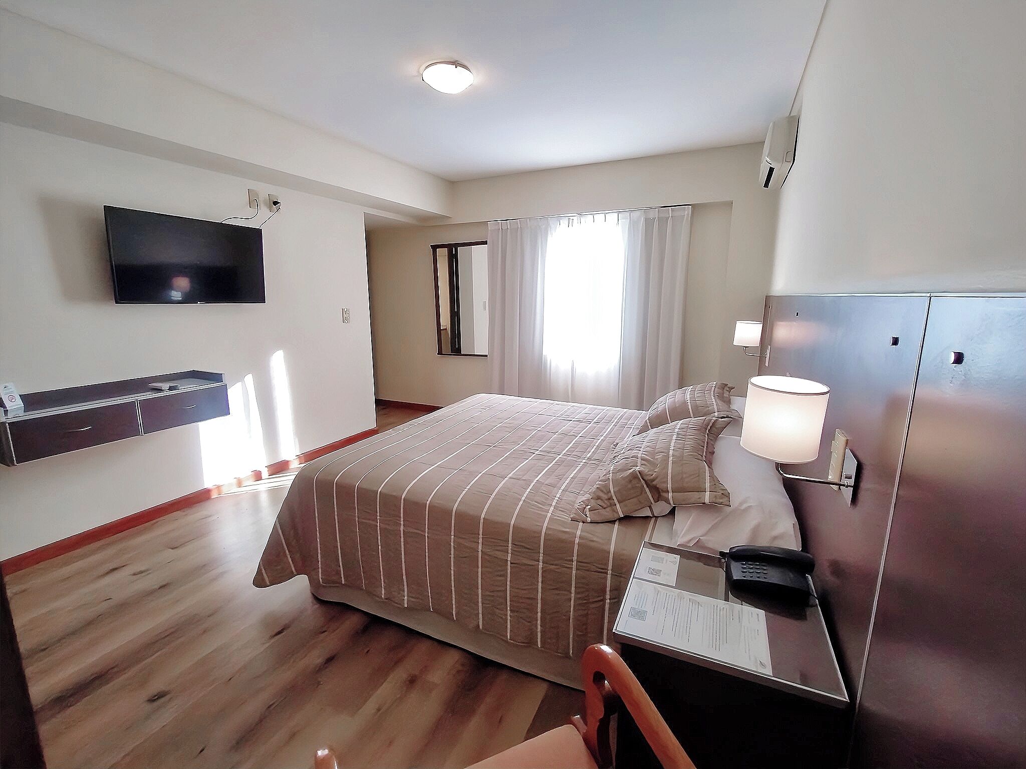 A hotel room with a bed, bedside table, lamp, and a TV mounted on the wall.