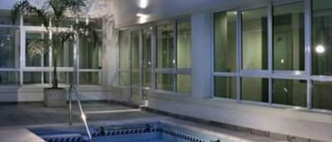 A swimming pool with a tiled deck, surrounded by a building with large windows and a palm tree.
